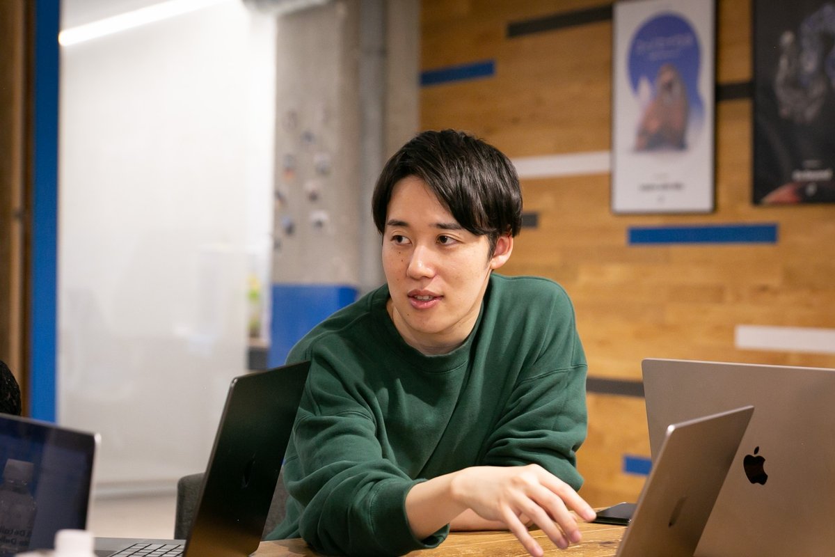男性のバストアップ写真。黒髪、黒い目。緑のトップス。顔は正面、目線はやや左を向いている。