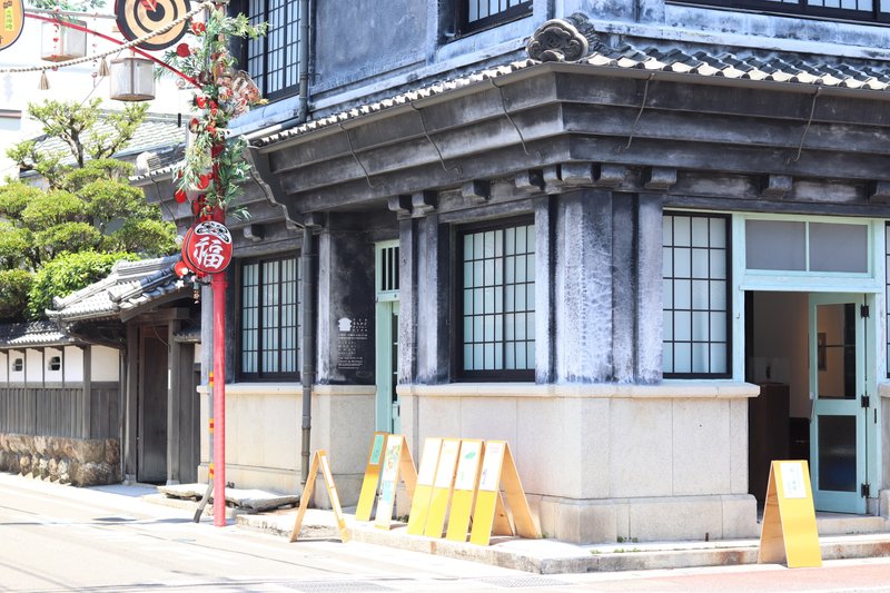 写真：通りの角に立つ古民家。立派な軒と漆喰の建築です。ギャラリーの前にはフライヤーを貼った立て看板が並んでいます。