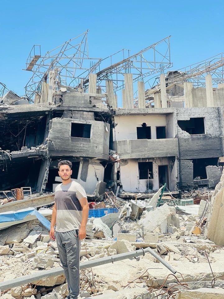爆撃され破壊された建物の前に立ち尽くすモハメド