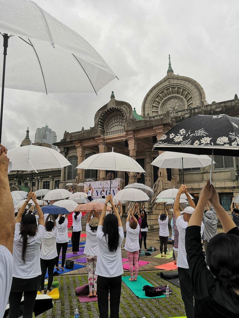 築地本願寺での第10回国際ヨガデーの感動的な瞬間:雨の中のヨガ 3 2024年6月21日、雨の築地本願寺で開催された第10回国際ヨガデーの様子。雨粒が地面を打つ音と共に、参加者たちは傘を差しながらヨガのポーズをとっています。