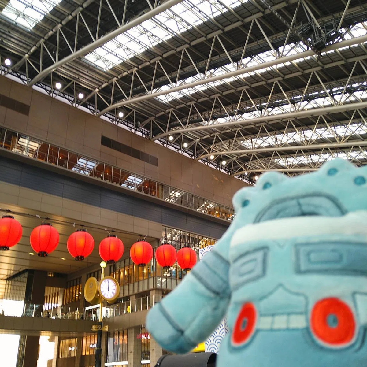 JR大阪駅の「時空の広場」で、駅の天井とルクアイーレの壁を見上げるドータクンのぬいぐるみの画像。夏にちなんで、広場には夏祭りのような赤い提灯の飾りつけがされている。