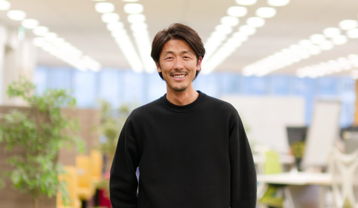 池端由基さんのバストショット。黒い長袖のセーターを着て、オフィスのような明るい室内で笑顔を見せている。背景にはデスクや椅子、観葉植物があり、天井の照明が奥に向かって並んでいる。