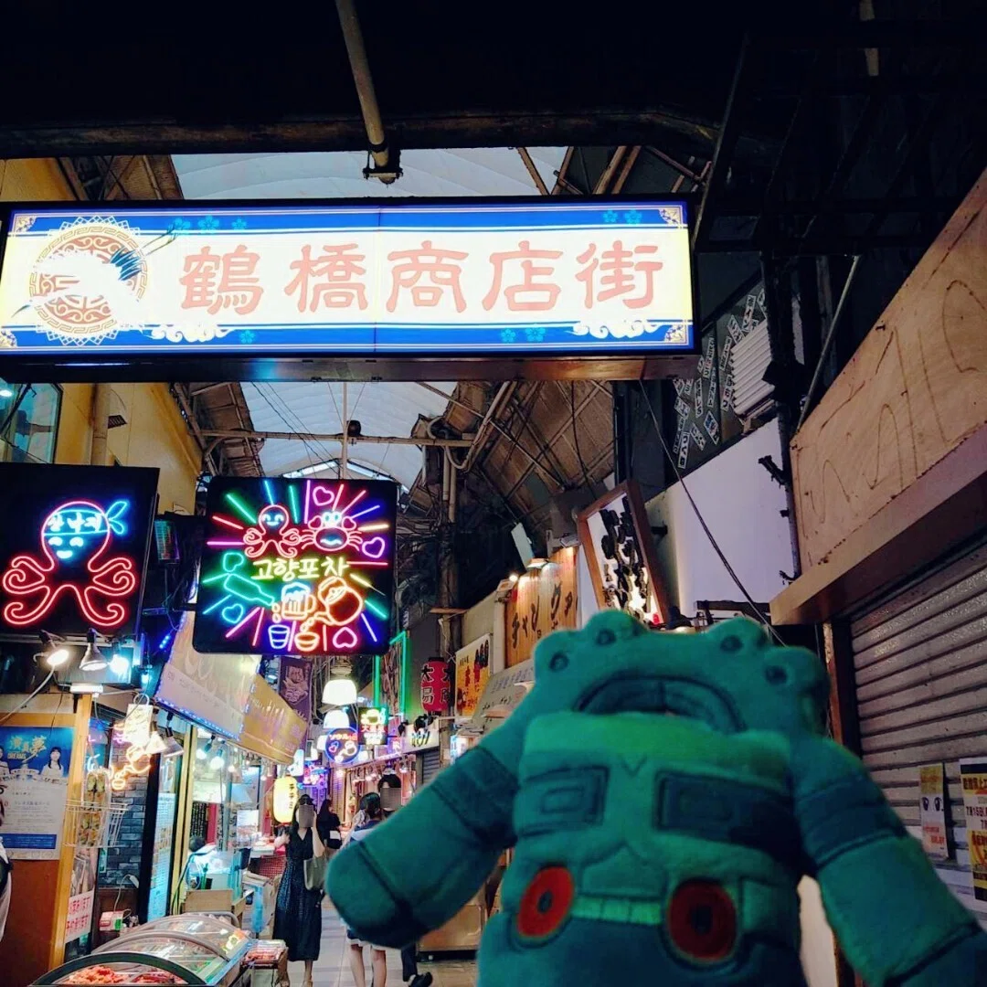 鶴橋商店街の画像。商店街の入口と、いくつかの商店、ネオンサインの看板、ドータクンのぬいぐるみが写っている。
