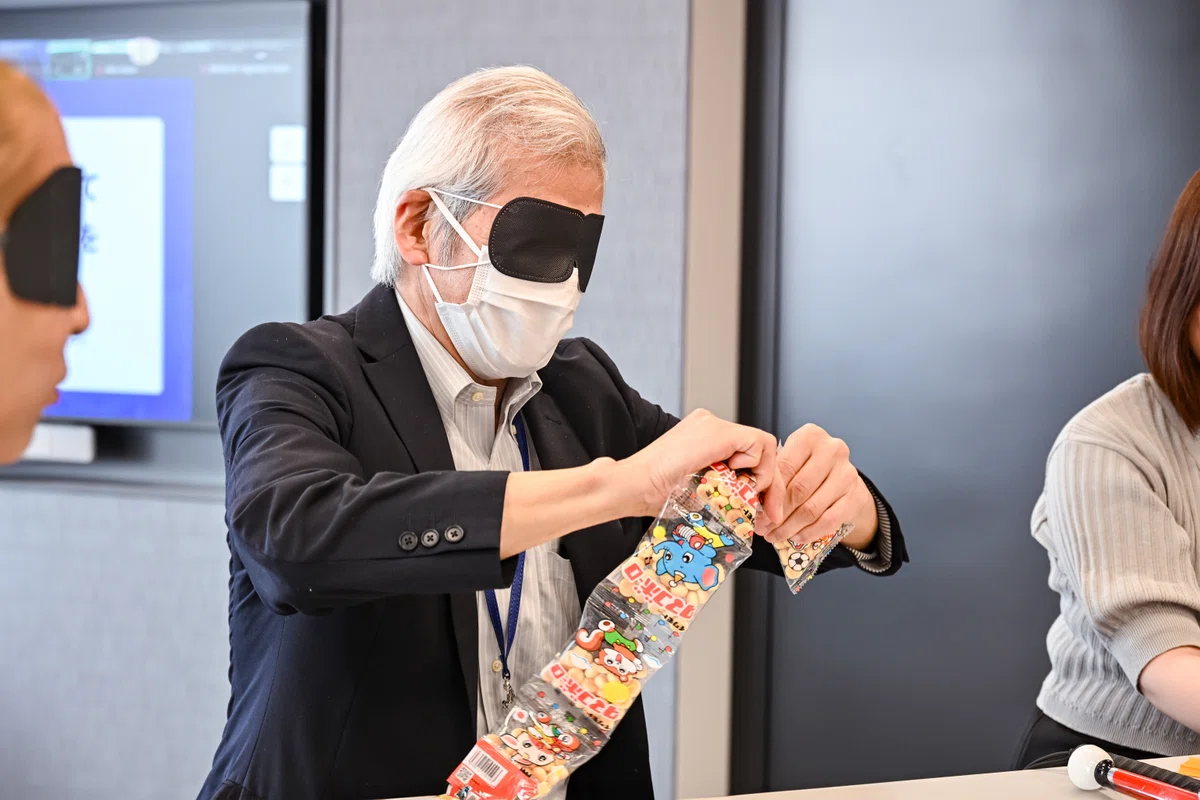 暗闇おやつのワークに取り組む男性の写真。アイマスクとマスクを着用し、手探りで５連卵ボーロの袋を切り分けようとしている。