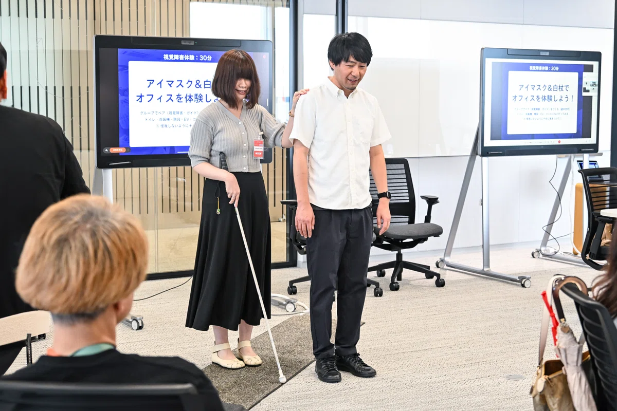 白杖の使い方とガイドの仕方をレクチャーするエガシラさんとタキザワ。エガシラさんは右手に白杖を持ち、左手でタキザワの右肩に乗せている。