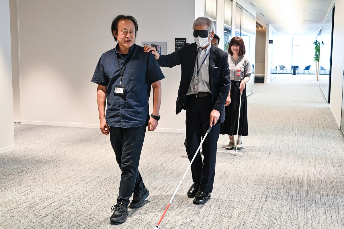参加者による白杖歩行体験の写真。アイマスクを着けて白杖を持った男性と、ガイド役の男性が並んで歩いている。