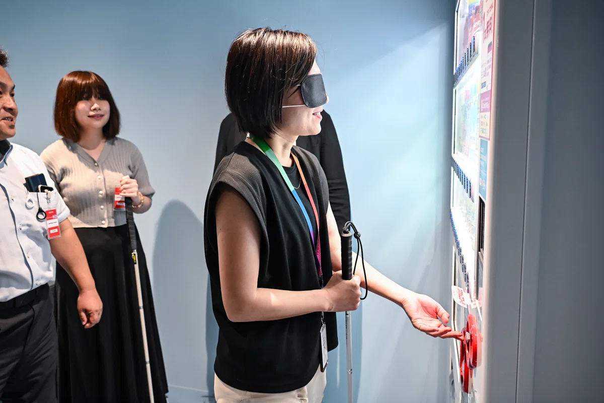 アイマスクをしたまま自動販売機を操作する女性の写真。右手に白杖を持ち、左手で自動販売機のボタンを手探りしている。