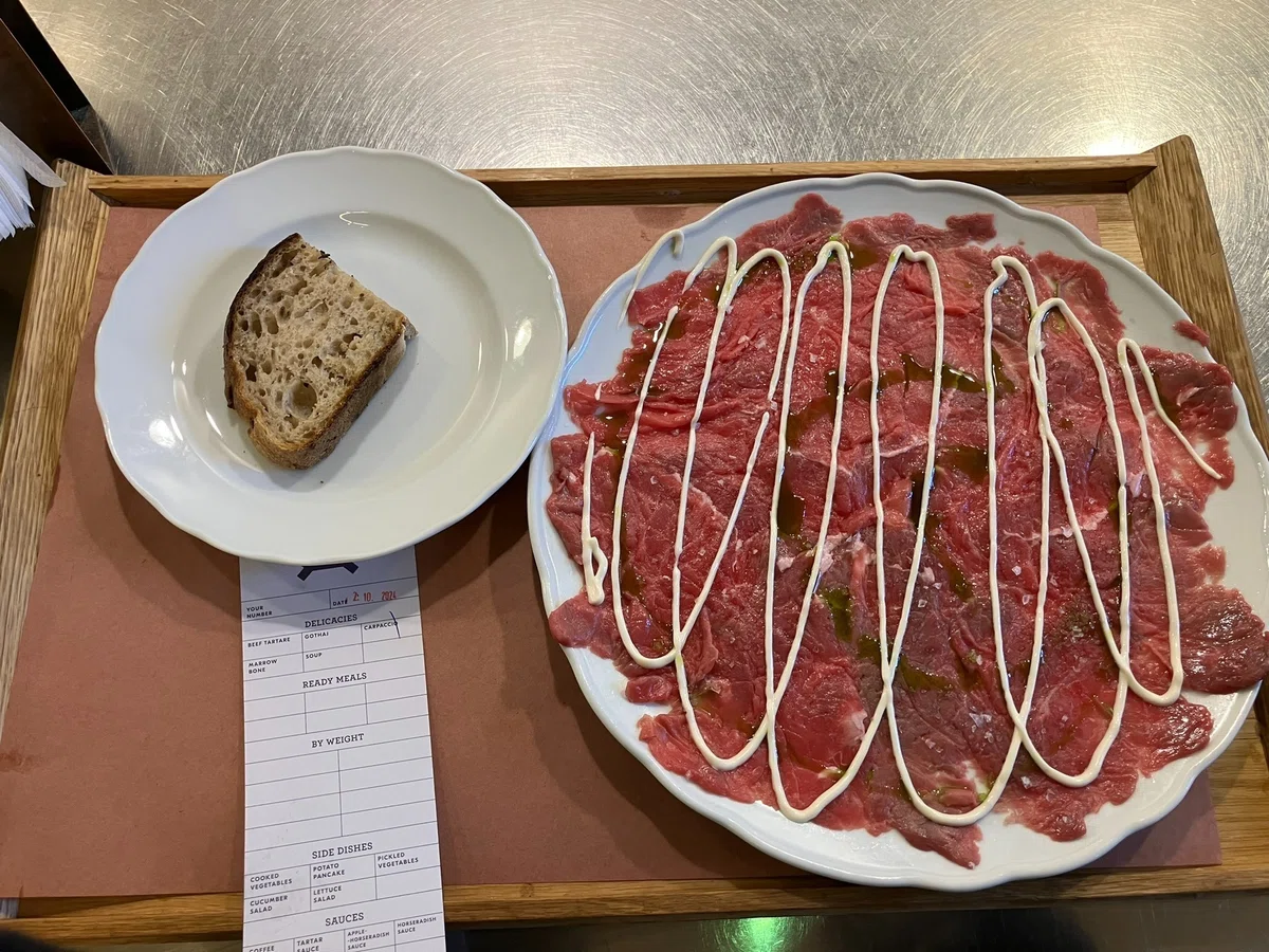 牛の薄切りの生肉に白いソースが掛かっている。左隣の小さい皿にはパンが載っている。