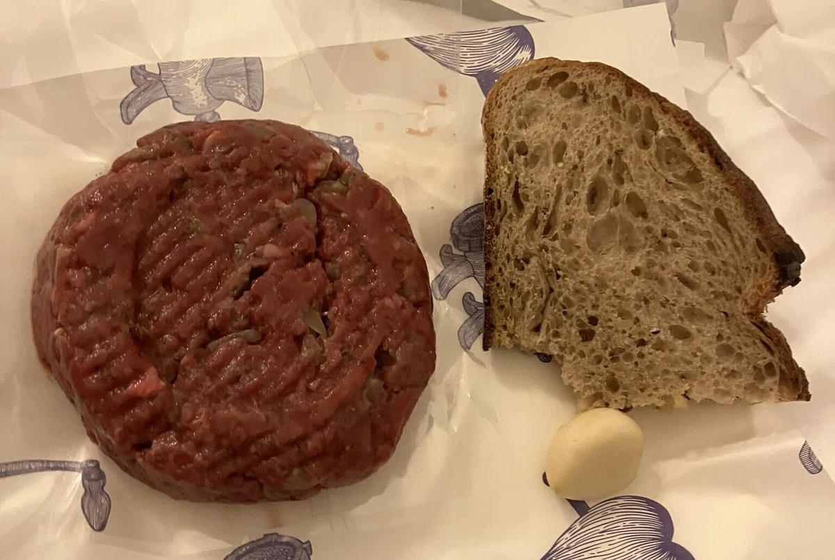 円形の生肉を叩いたものと右隣にパン一切れとにんにくひとかけ。