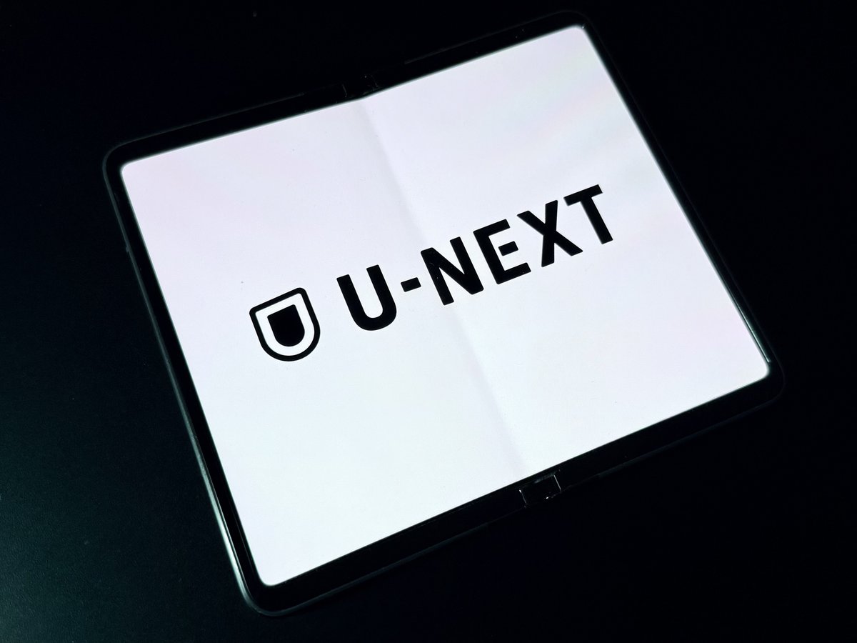 折りたたみスマホの画面に「U-NEXT」のロゴが大きく表示されています。画面は白い背景に黒いロゴが配置されており、シンプルで洗練されたデザインです。タブレットの背景は黒一色で、画面の明るさが際立っています。