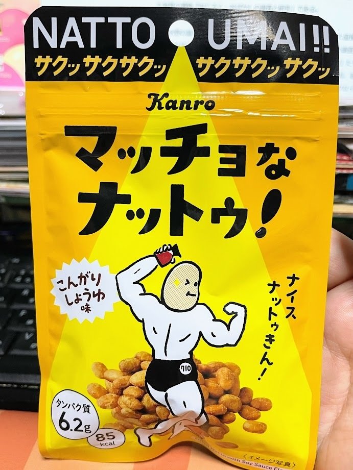 Kanro「マッチョなナットゥ！」のパッケージ　黄色い背景にマッチョな肉体のついた大豆が醤油を持ってマッチョポーズをとっているイラスト　「ナイスナットゥきん！」　こんがりしょうゆ味　タンパク質6.2g　85kcal