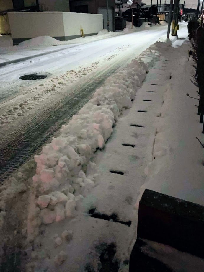 夜の道　車道は除雪されており、脇に堤のように雪が積み上がっている　歩道部分は平らだが側溝の蓋の穴が見える