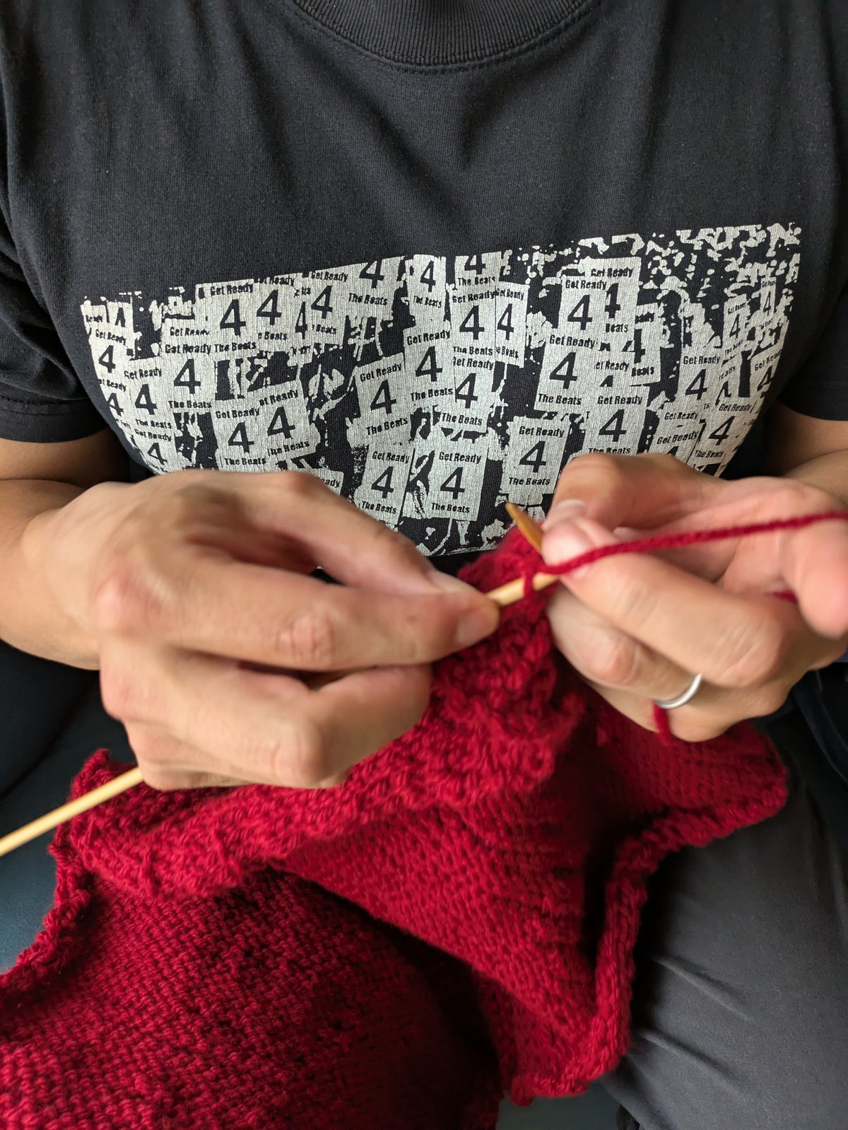 Her thick fingers are manipulating knitting needles on the front of her black T-shirt, knitting a piece of red yarn.