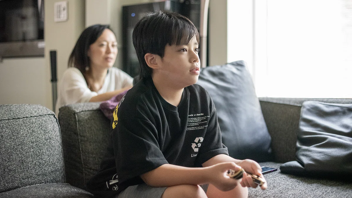 Switchのコントローラーを持ち、真剣な表情でゲームに取り組む小学生の男の子。後ろには見守る母親の姿。