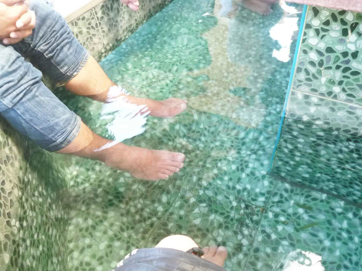 タイル張りの足湯に大人の足が浸かっているクローズアップ。縁に座る人の膝や足先が見え、水面がゆるく揺れている。