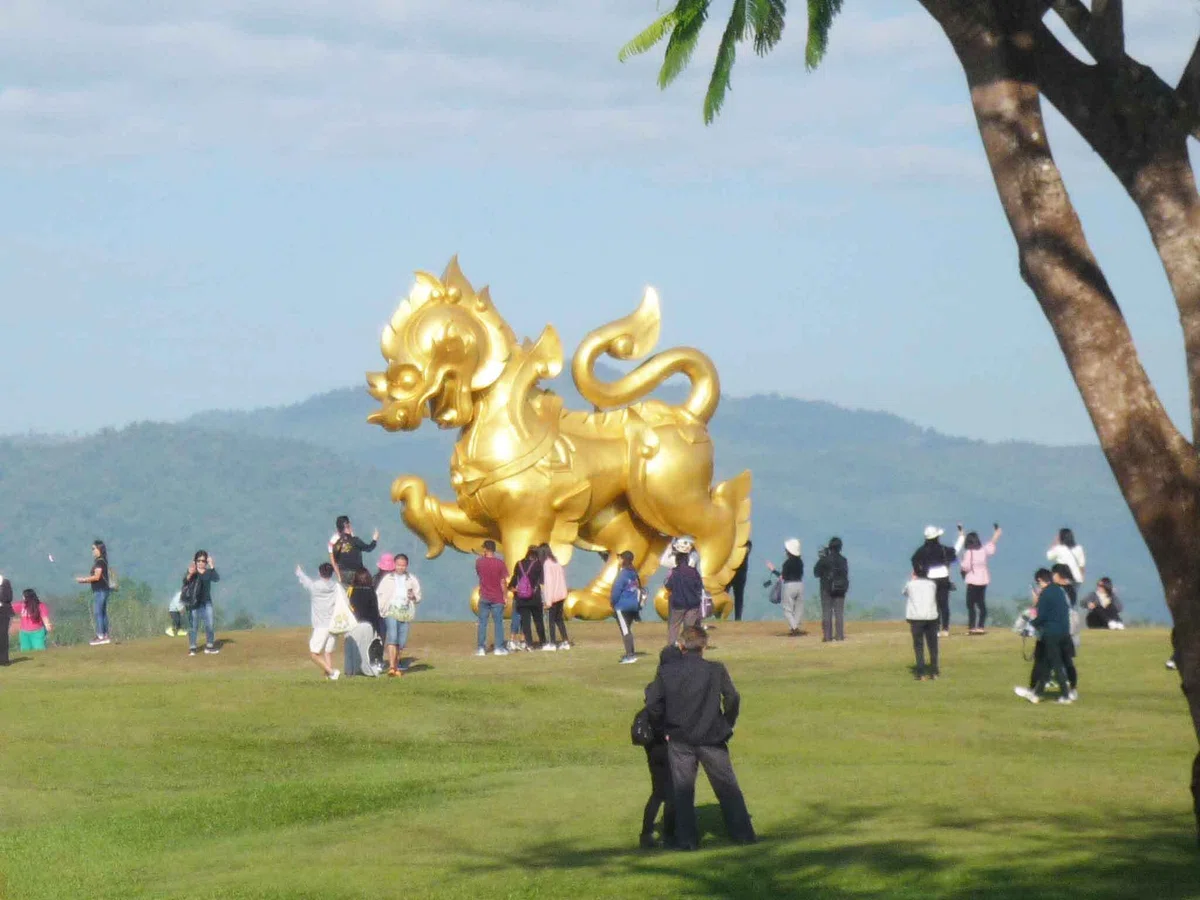 芝生の丘の中央に金色のシンハ像。周囲で人々が記念撮影し、背後に山並みが見える。