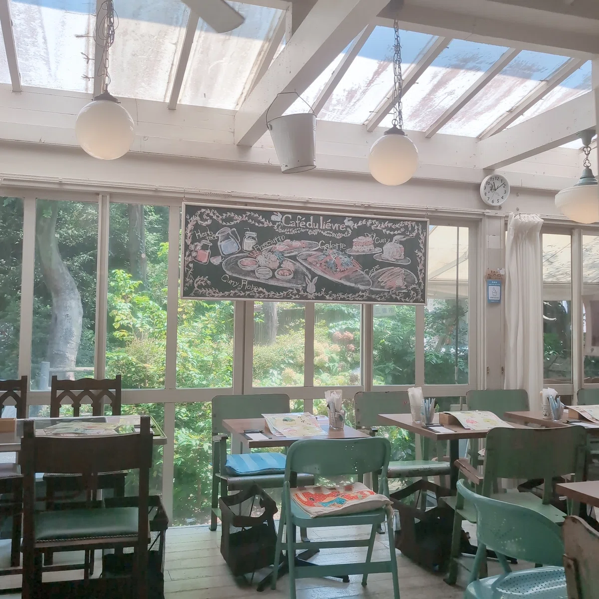 カフェの店内。ガラスの天井窓から陽の光が差し込む温かな雰囲気。中央にはチョークアートの施された黒板があり、緑の椅子や木の椅子の置かれたテーブル席が並ぶ。