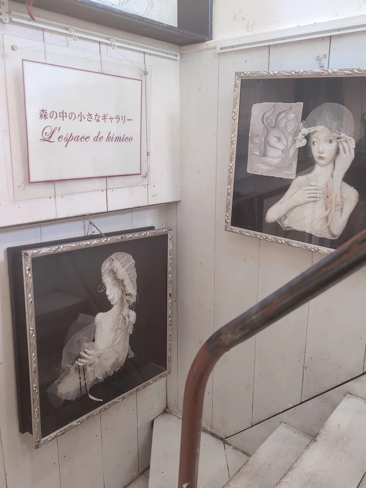 森の中の小さなギャラリーと書かれた看板と絵画がかかる壁。少女の絵や人形がかけられている