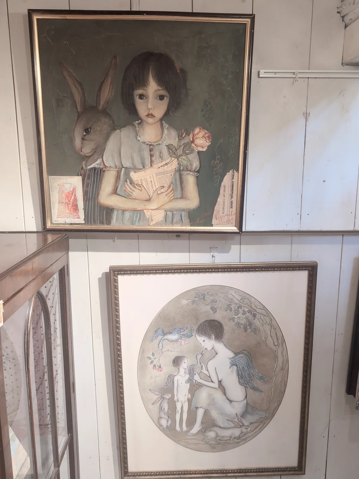白い木製の壁にかけられた絵画。うさぎやバラを持った少女、天使の羽を持つ二人の人物などが描かれている