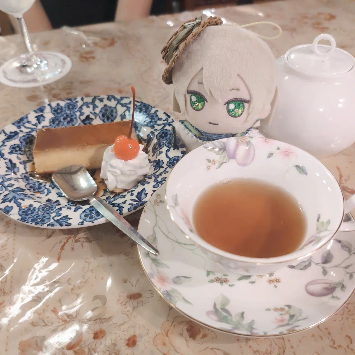 カフェのテーブル。紅茶の注がれた花模様のカップアンドソーサー、さくらんぼとホイップクリームが添えられた横長のプリン。