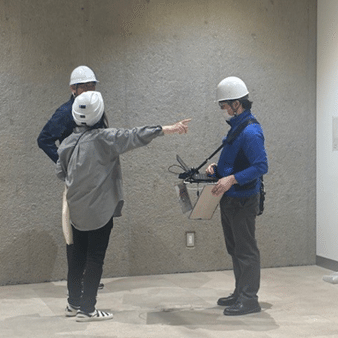 施設職員１名（写真左、白いヘルメットを被り、上着に灰色の作業着を着ている）と通信事業者の専門作業員２名（写真右と施設職員の後ろ、白いヘルメットを被り、上着に青色の作業着を着ている）が、ミュージアムショップとして整備される予定の区域で電波測定の結果について話し合いを重ねている様子。