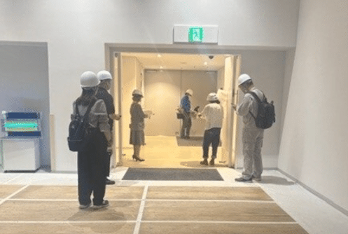 非常口の奥で、機器を手に電波測定している通信事業者の専門作業員（白いヘルメットを被り、上着に青色の作業着を着ている）と、非常口付近及び手前で電波測定に同行している施設の職員と東京都職員。