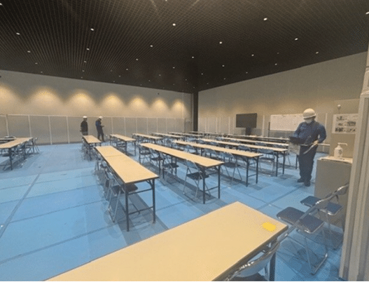 室内の縦２列、横５列に並べられたベージュの長机付近（床はブルーシート）で通信事業者の専門作業員（写真右、白いヘルメットを被り、上着に青色の作業着を着ている）が機器を手に電波測定している様子