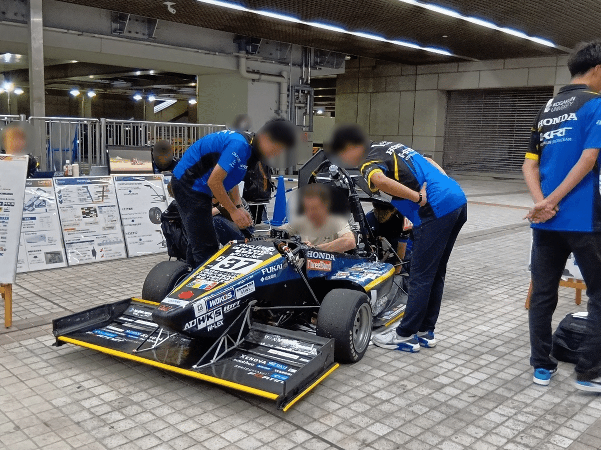 地元大学の学生がフォーミュラカー(黒色。様々な企業のロゴが入っている。)について説明し、来場者が車両に乗り込みながら聞いている。