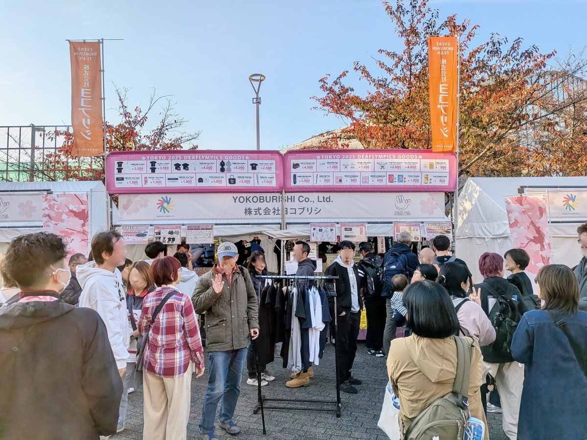 グッズ販売ブース。たくさんの人が列を作っている。店先にはシャツなどの見本がラックにかかっている