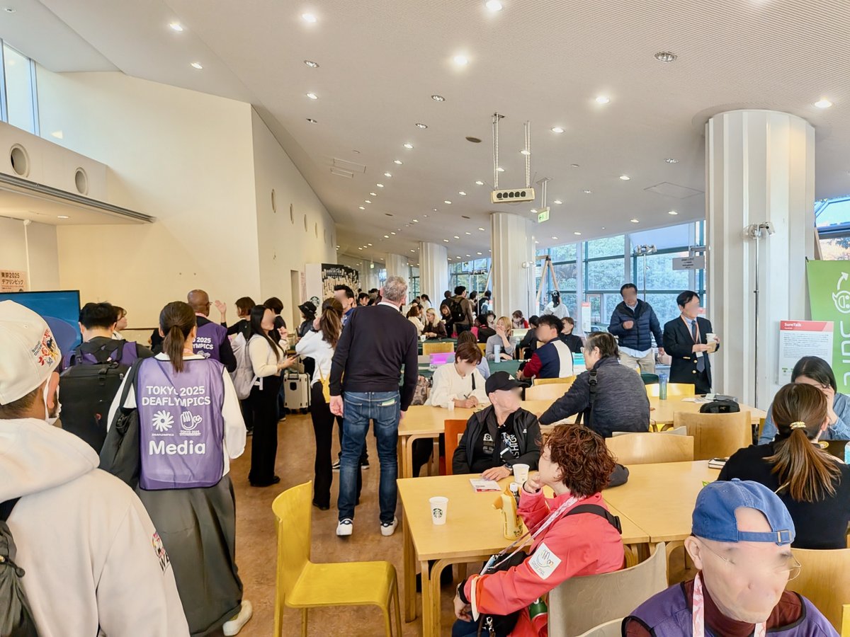 カフェスペースの様子。社食や学食のような、白い空間に木目調のテーブルが並んでいる。どれも満席。一般客だけでなくスタッフゼッケンを着けた人や海外選手と思われる人たちも交流していた