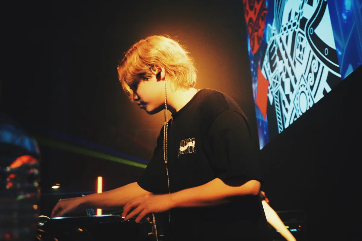 渋谷WOMBのステージでDJ KENDIが集中してプレイする様子。背後に照明とNESTALのアートビジュアルが映り、黄金色のライトが印象的。
A DJ KENDI performing with full focus at WOMB Shibuya, illuminated by golden light and the signature NESTAL visual backdrop.