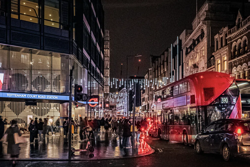 夜のロンドン、トッテナム・コート・ロード駅前の風景。雨に濡れた石畳のような路面が、街灯やネオン、そして赤い二階建てバスのテールランプを鏡のように反射している。左側には「TOTTENHAM COURT ROAD STATION」と書かれたモダンで明るい地下鉄駅の入り口があり、多くの人々が行き交う都会の躍動感が伝わってくる。