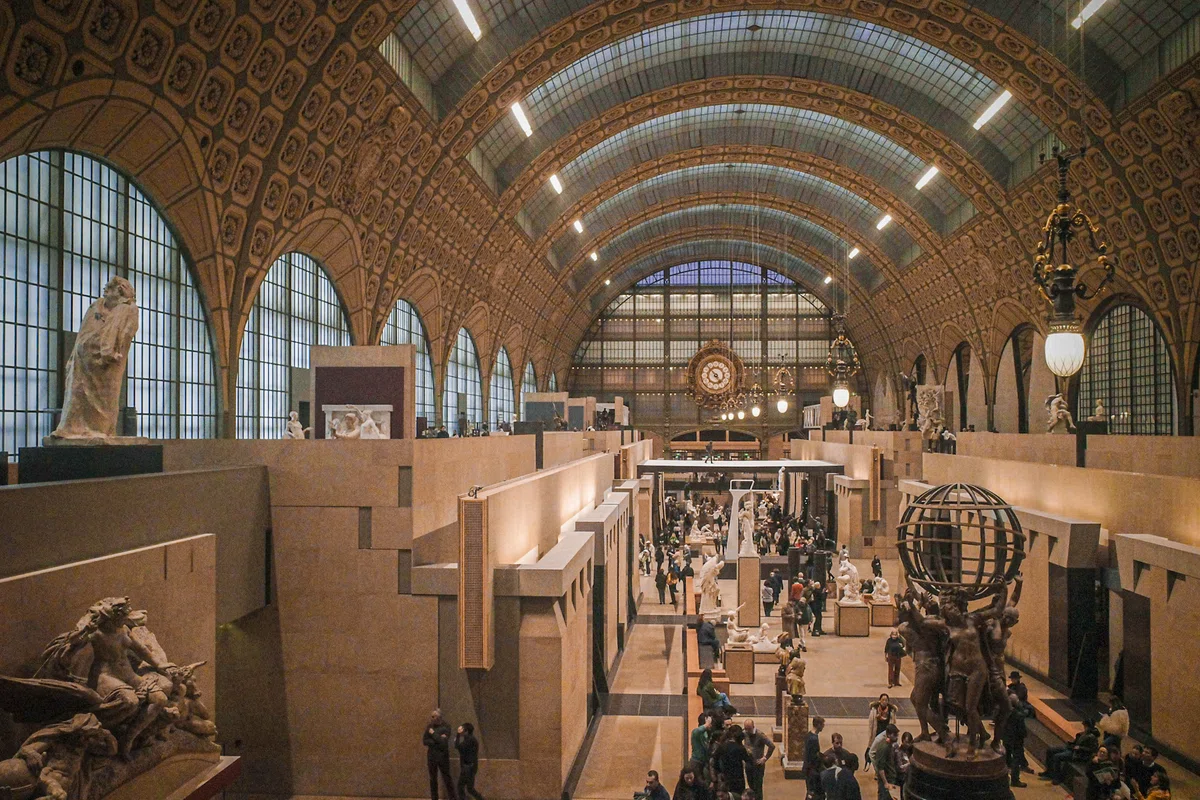 パリ・オルセー美術館のメインホール。かつての駅舎の面影を残す巨大なアーチ状のガラス天井と、奥に見える象徴的な大時計、そして中央通路に並ぶ彫刻群が織りなす美しい館内の風景。