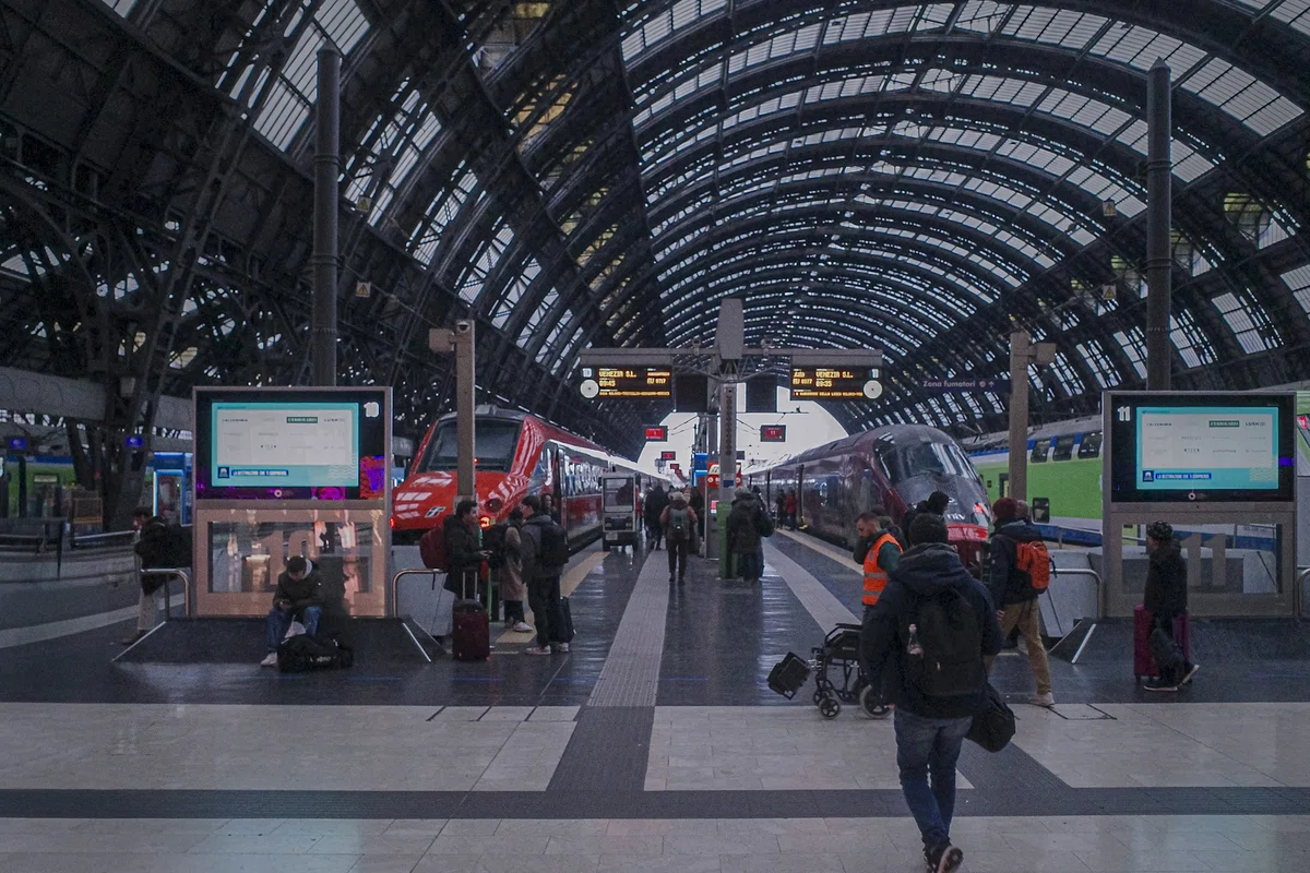 イタリア・ミラノ中央駅の巨大なプラットホーム。鉄骨とガラスで組まれた高いアーチ屋根の下、停車中の列車やスーツケースを持った旅行者たちが行き交う、ヨーロッパの旅情あふれる光景。
