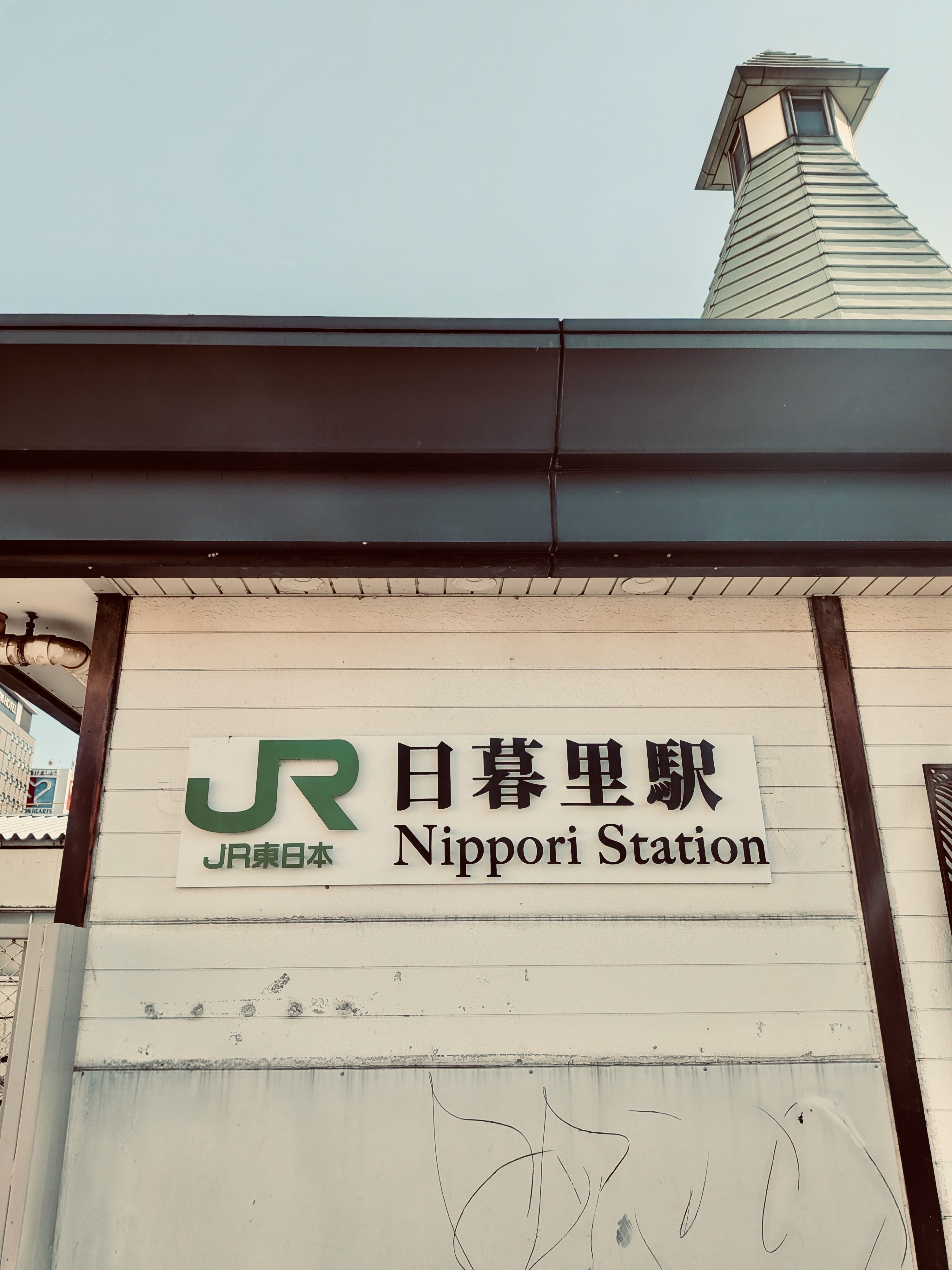 JR日暮里駅。日暮里駅という看板。その屋根の向こうに、物見櫓のようなものがたっている