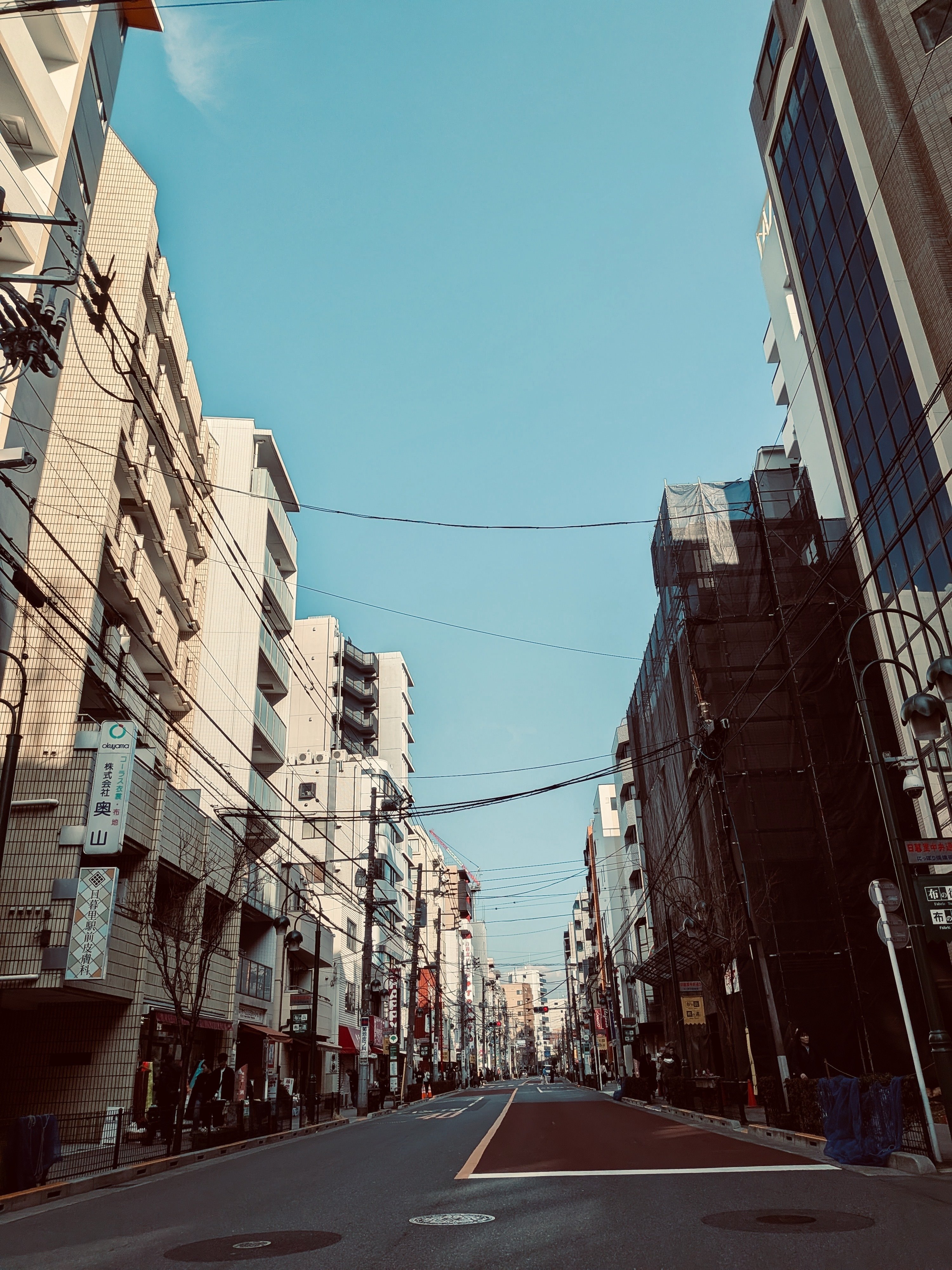 日暮里の街並み。道の両側に高くない殺風景なビルが立ち並び、道を跨いで電線が蜘蛛の巣のようにかかっている。ビルの向こうには青空。