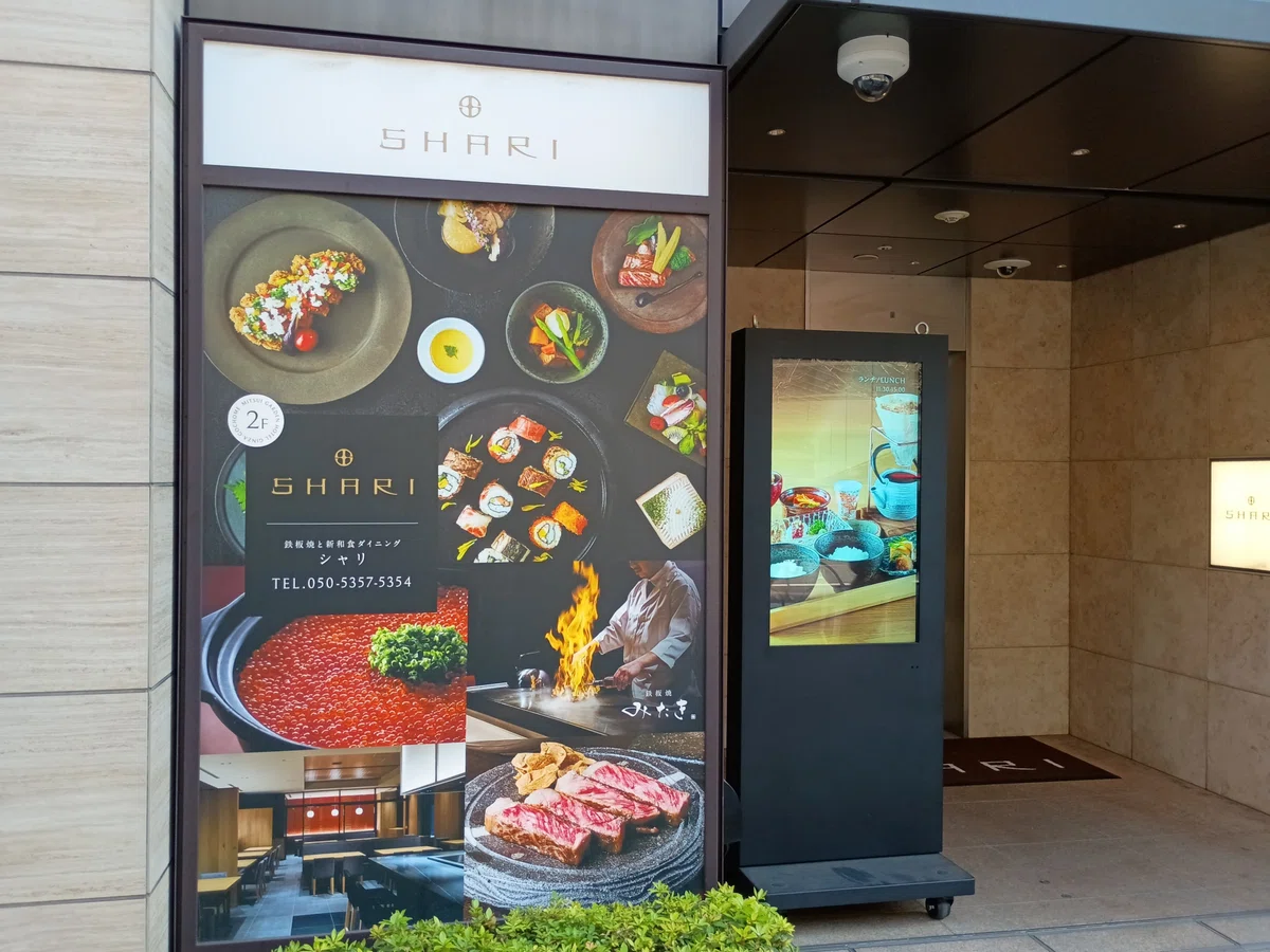 （看板の写真）三井ガーデンホテル銀座五丁目の1階にある、鉄板焼と新和食ダイニング「SHARI」の立て看板と入り口。看板には美味しそうな鉄板焼きやロール寿司などの写真がデザインされている。