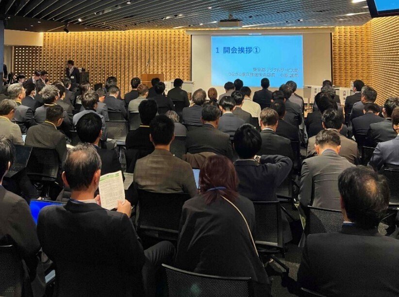 説明会会場に、スーツを着用した約80名が集まり、正面に設置された説明会用の投影スクリーンに向かって着席している様子。