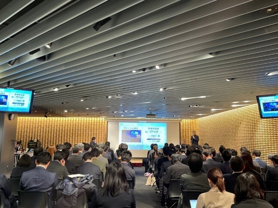 ジャーナリストの石川 温氏がモデレーターを務め、KDDI株式会社およびソフトバンク株式会社の登壇者が、説明会会場の登壇スペースで発表を行っている様子。会場には約80名の参加者が着席し、登壇者の説明に耳を傾けている。