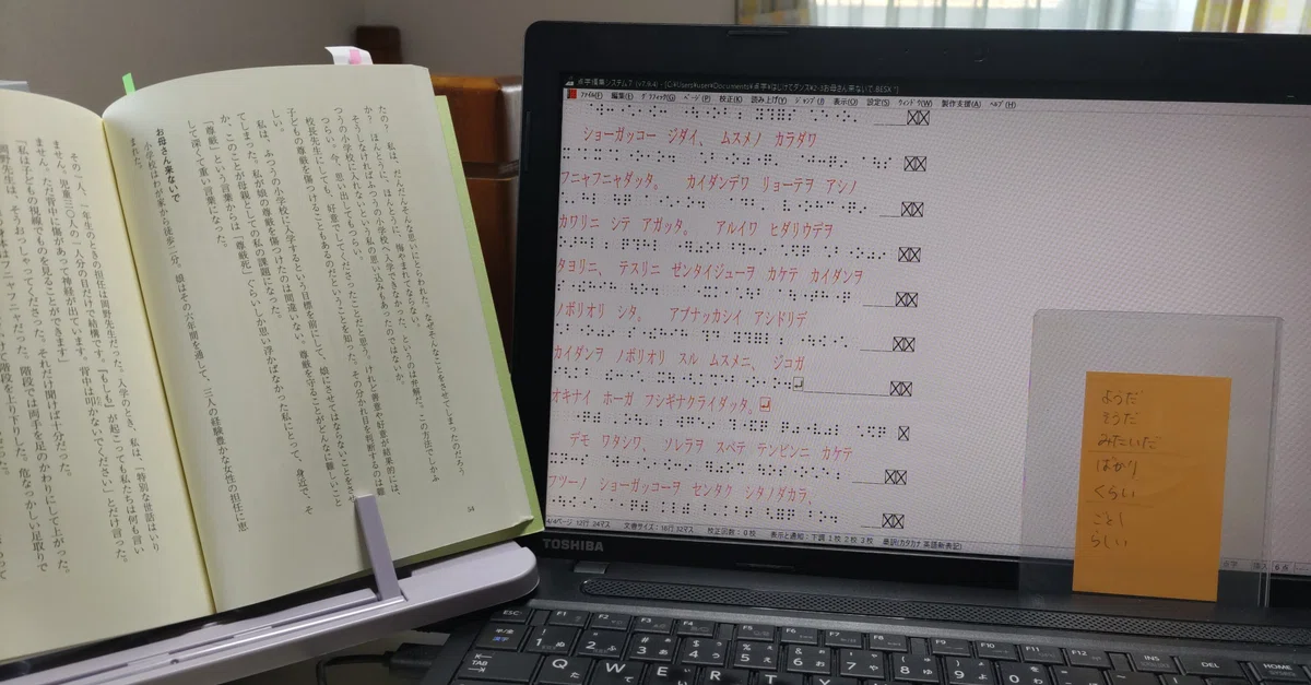 ノートパソコンの画面左寄りに点訳データが表示され、右側の空白部分にオレンジ色のカードが立てかけられている。ノートパソコンの左脇には百均の読書台に立てられた点訳中の書籍『はじけてダンス！』。