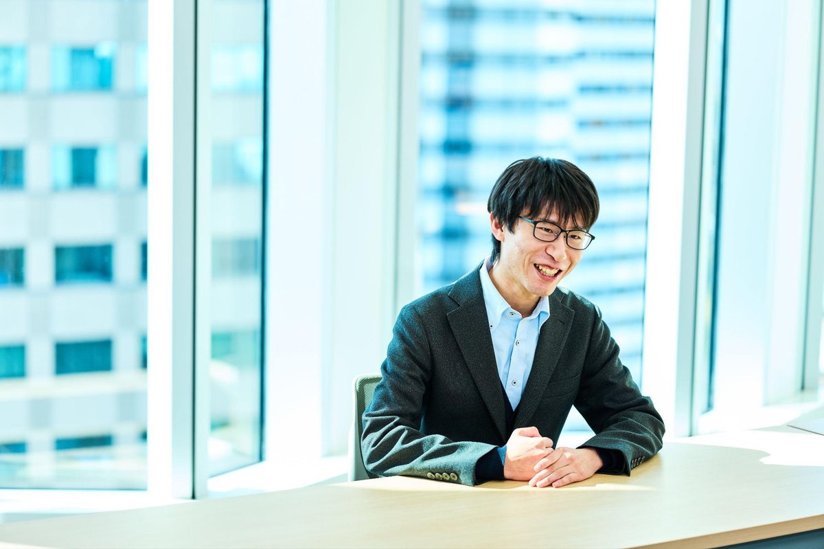 明るいオフィスの会議室で、黒縁の眼鏡をかけた男性(佐々木)が、グレーのジャケットと水色のシャツを着て、木製のテーブルに手を組んで座っている。背景には大きなガラス窓があり、都市のビル群が見える。柔らかな自然光が差し込み、明るく爽やかな雰囲気。男性は穏やかな笑顔でカメラを見ている