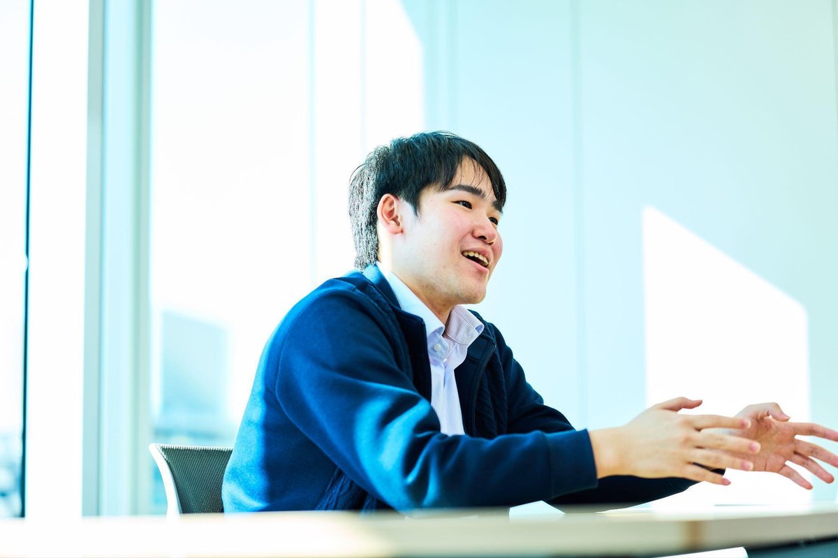 明るいオフィスで、紺色のジャケットと水色のシャツを着たの男性(本田)が、白いテーブルに座り、両手を前に出して身振りを交えながら話している。表情は明るく、笑顔で会話に集中している様子。背景には大きなガラス窓があり、都市の景色が見える