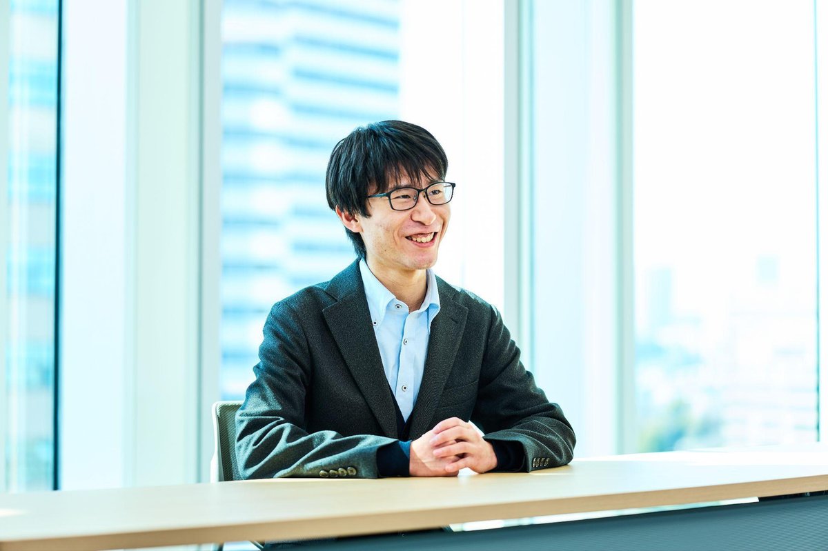 明るいオフィスの会議室で、黒縁の眼鏡をかけた男性(佐々木)が、グレーのジャケットと水色のシャツを着て、木製のテーブルに手を組んで座っている。穏やかな笑顔でカメラを見つめており、リラックスした雰囲気。背景には大きなガラス窓があり、都市のビル群が見える