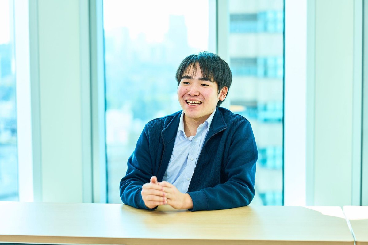 明るいオフィスで、紺色のジャケットと水色のシャツを着た男性(本田)が、木製のテーブルに手を組んで座り、満面の笑みを浮かべている。表情は非常に明るく、楽しそうな雰囲気。背景には大きなガラス窓があり、都市のビル群が見える。全体に明るく開放的な印象