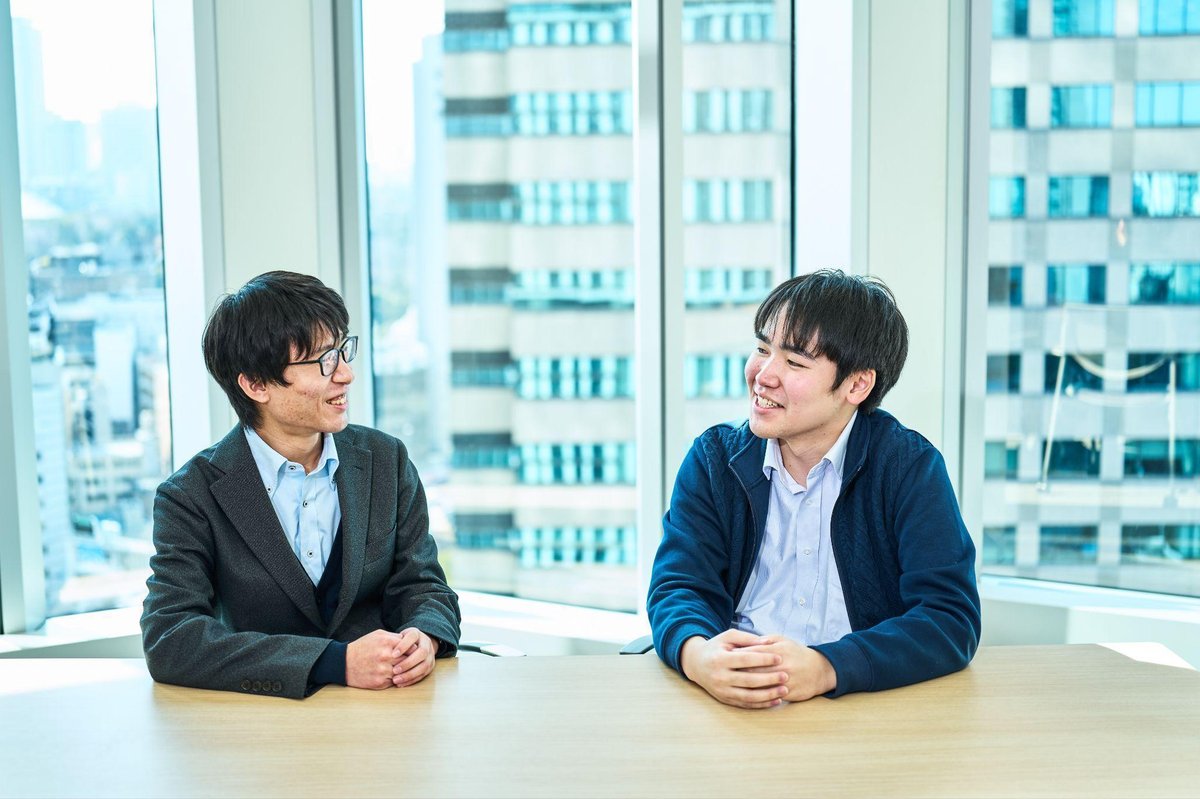 明るいオフィスの会議室で、2人の男性が木製のテーブルを挟んで向かい合って座り、笑顔で会話している。左側の男性(佐々木)は黒縁の眼鏡をかけ、グレーのジャケットと水色のシャツを着用。右側の男性(本田)は紺色のジャケットと水色のシャツを着用。両者とも手をテーブルに置き、リラックスした雰囲気で互いを見つめている。背景には大きなガラス窓があり、都市のビル群が見える