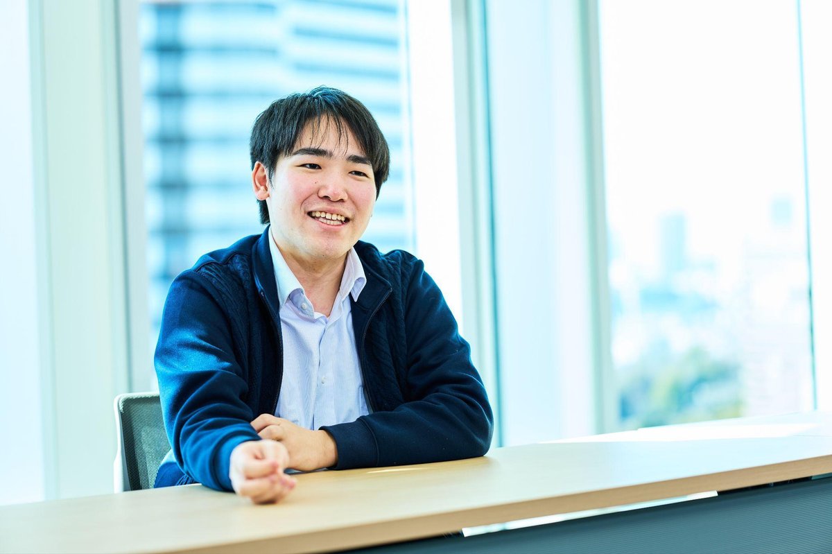 明るいオフィスで、紺色のジャケットと水色のシャツを着た男性(本田)が、木製のテーブルに座り、笑顔でカメラを見ている。手はテーブルの上に置かれており、リラックスした姿勢。背景には大きなガラス窓があり、都市の景色が見える。全体に明るく爽やかな雰囲気