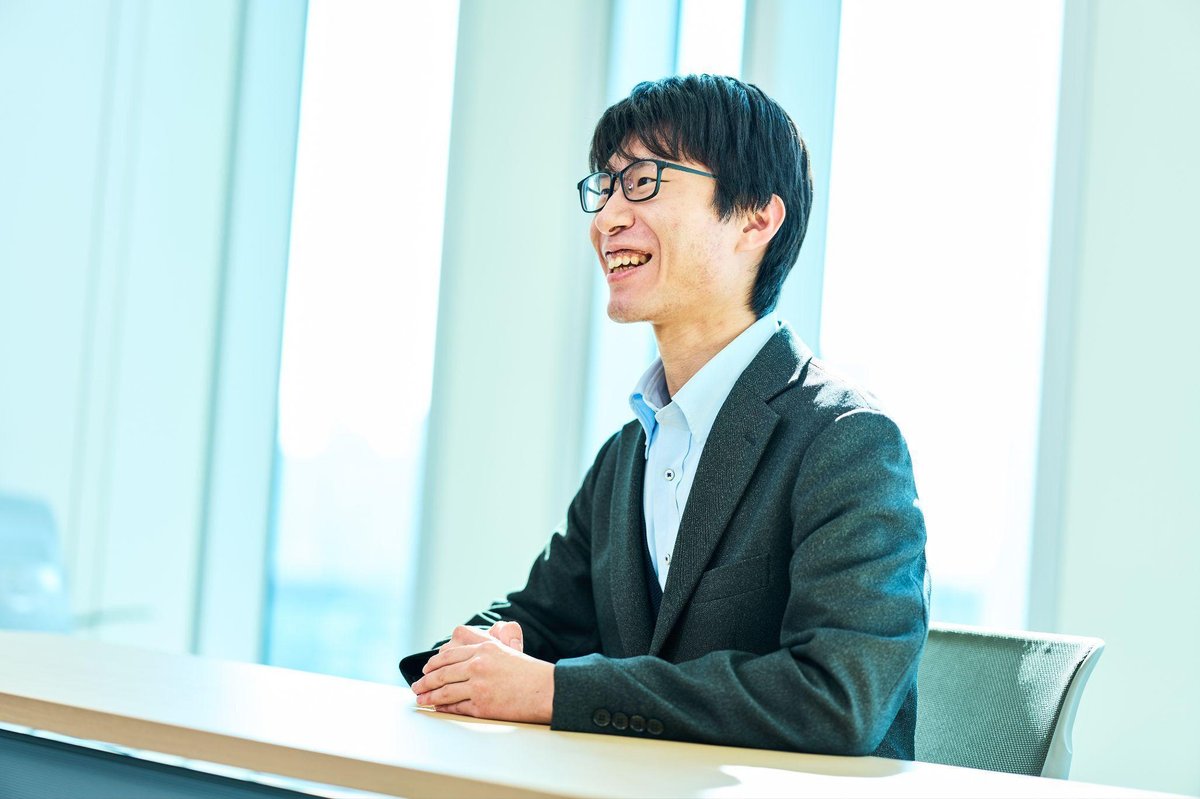 明るいオフィスで、黒縁の眼鏡をかけた男性(佐々木)が、グレーのジャケットと水色のシャツを着て、木製のテーブルに手を組んで座っている。顔を左斜め上に向けて、明るい笑顔を浮かべている。背景には大きなガラス窓があり、柔らかな自然光が差し込んでいる。全体に明るく開放的な雰囲気