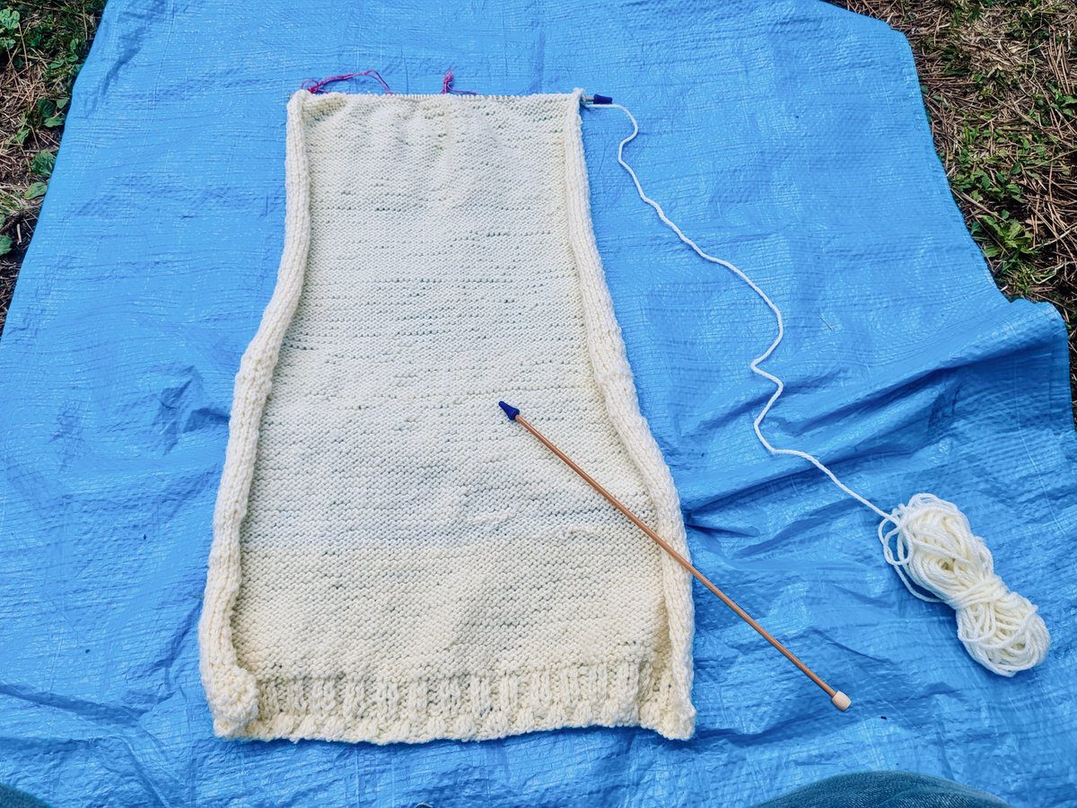 セーターの編み地。前半分。ブルーシートの上に、白い編み地が載っている。編み棒にはブルーのキャップがはめられている