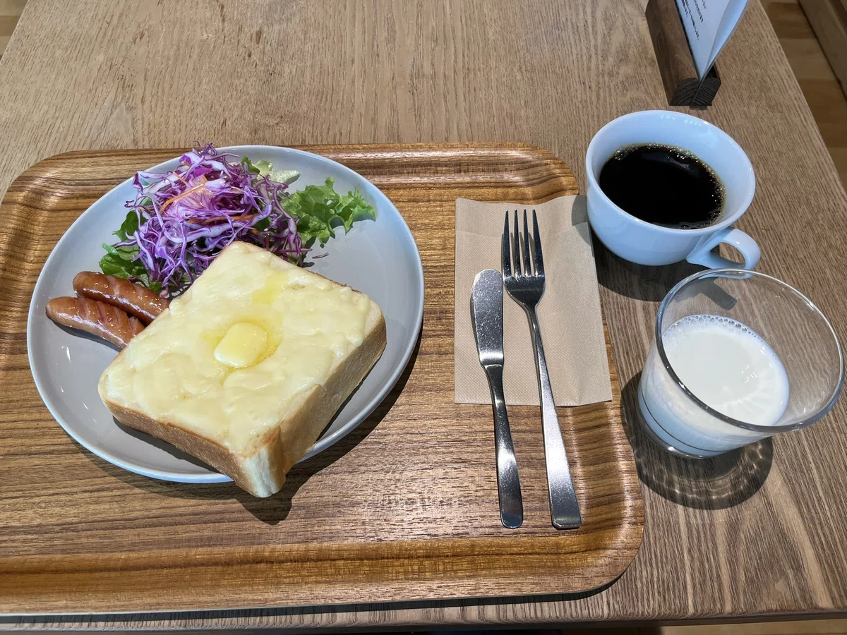 木のトレイの上の大きめの皿に厚切りチーズトースト、ウィンナー2本、サラダが盛り付けられている。バターナイフとフォークが、紙ナプキンの上に乗っている。トレイの横に、コーヒーと牛乳のグラスが置かれている