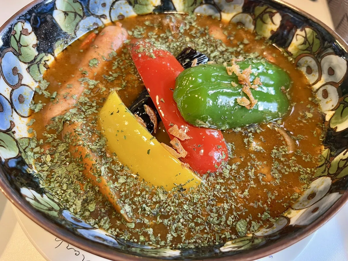 魔女のスプーンの薬膳カレー。ピーマン、パプリカ（赤、黄）、なす、ウインナーがスープに入っている。緑色のハーブが、たっぷりかかっている。